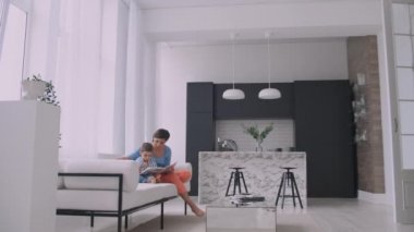Kanepede oturma odasında evin parlak beyaz iç oturan bir kitap okuma bir çocuk ile genç bir anne.