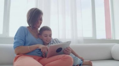Anne ve oğlu dijital tablet kullanarak koltukta oturuyor. Tablet dokunmatik ekranlı bir video izlerken birlikte mutlu anne ve küçük çocuk kullanarak. Gülümseyen anne ve sevimli çocuk dijital tablet üzerinde oynama