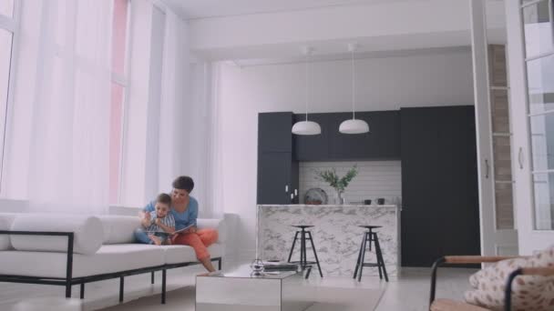 Mladá matka s dítětem, která čte knížku v jasně bílém interiéru domu v obývacím pokoji na gauči. — Stock video