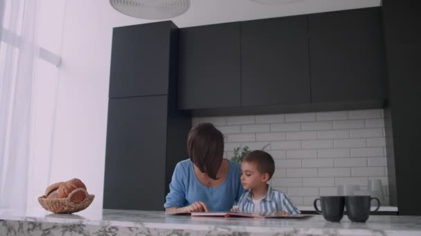 Young Spanish mother and son sitting at the table reading a book together and smiling. — Stock Video