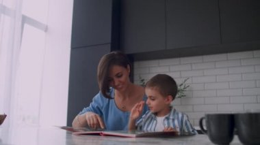 Genç güzel anne ve oğlu bir kitap okuma ve bir kitap içine bir parmak alay resimlere bakarak ve sayfalarda leafing parlak bir mutfakta bir masada oturan.