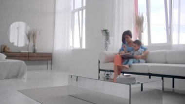 Kanepede oturma odasında evin parlak beyaz iç oturan bir kitap okuma bir çocuk ile genç bir anne.