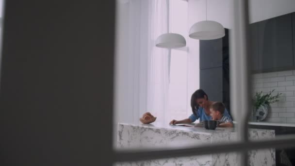 Eine junge spanische Mutter mit ihrem Sohn, der am Tisch sitzt, bringt dem Kind das Lesen bei und fordert seinen Sohn auf, ihm zu helfen, — Stockvideo
