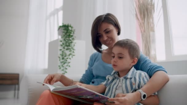 Una madre joven con un niño leyendo un libro sentado en un interior blanco brillante de la casa en la sala de estar en el sofá . — Vídeos de Stock