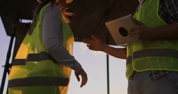 タブレットを持ったビルダーの男性と白いヘルメットをかぶった女性検査官は、建物の屋根の上に立って日没時に握手をします。成功した仕事の合意の象徴. — ストック動画