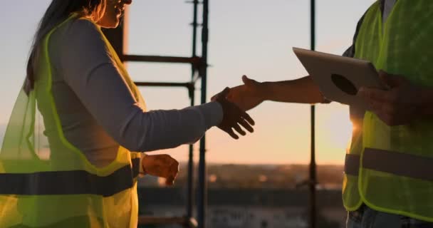 Bouwers man met een Tablet en een vrouw in witte helmen schudden handen bij zonsondergang staan op het dak van het gebouw. — Stockvideo