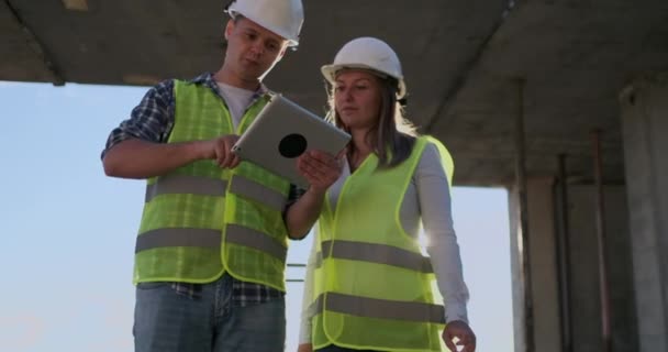 Bauarbeiter Mann und Architektin mit Helm, besprechen den Plan für den Bau eines Hauses, erzählen einander von dem Entwurf, halten ein Tablet in der Hand, betrachten die Zeichnungen, Hintergrund der Sonnenstrahlen — Stockvideo