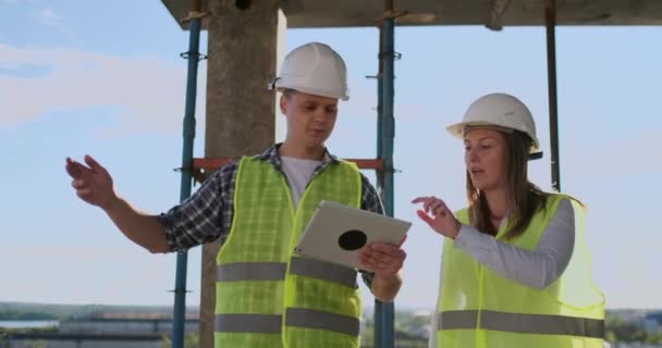 Travailleur de la construction homme et femme architecte dans un casque, discuter du plan de construction de la maison, parler de la conception, tenant une tablette, regarder les dessins, fond des rayons du soleil — Video