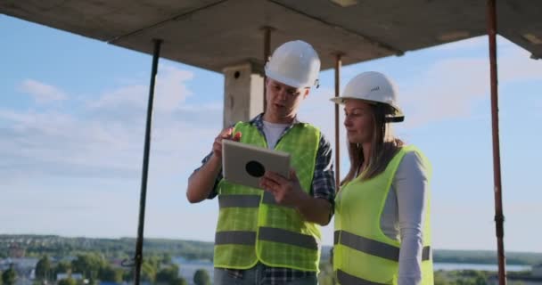 Byggande arbetare man och arkitekt kvinna i en hjälm, diskutera planen för byggandet av hus, berätta för varandra om designen, håller en tablett, titta på ritningar, bakgrund av solens strålar — Stockvideo