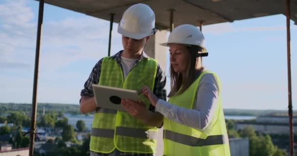 Bauarbeiter Mann und Architektin mit Helm, besprechen den Plan für den Bau eines Hauses, erzählen einander von dem Entwurf, halten ein Tablet in der Hand, betrachten die Zeichnungen, Hintergrund der Sonnenstrahlen — Stockvideo