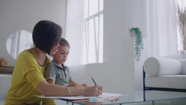 Mère en veste jaune et fils en t-shirt s'assoient à la table et dessinent ensemble des crayons de couleur sur papier. Bonne enfance. Aimer aide son fils dans la formation préscolaire et développe la créativité chez l'enfant . — Video