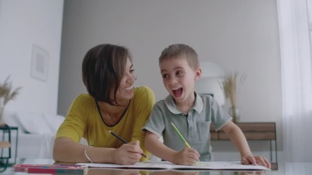Bella giovane madre in un maglione caldo sdraiato sul pavimento con mio figlio che disegna con pennarelli su carta raffiguranti la sua famiglia. Il bambino impara a disegnare . — Video Stock