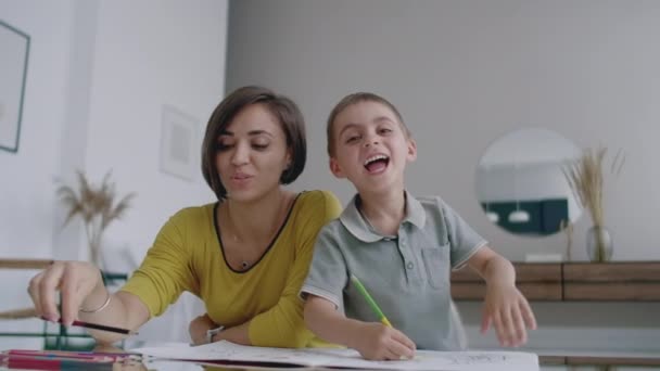 Mor i gul jacka och son i t-shirt sitta vid bordet och rita tillsammans färgpennor på papper. Lycklig barndom. Loving hjälper sin son i för skolan utbildning och utvecklar kreativitet i barnet. — Stockvideo