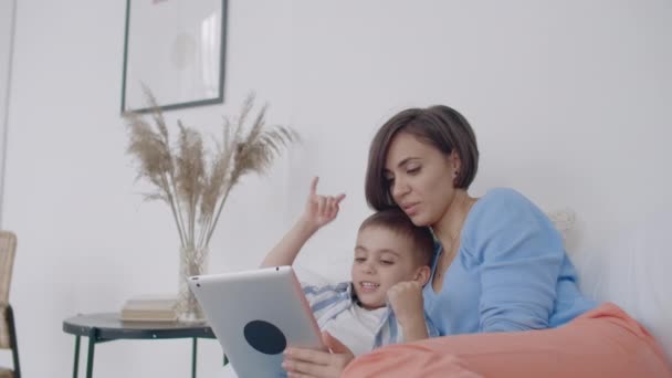 Mutter und Sohn schauen auf den Tablet-Bildschirm, der auf einem weißen Bett liegt. Spiele mit deinem Sohn auf deinem Tablet-Computer und schaue lustige Videos — Stockvideo