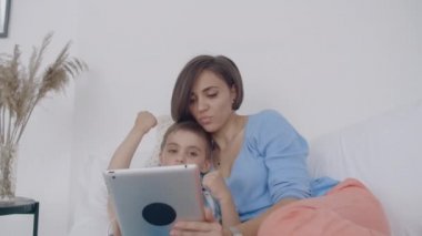 Beyaz yatak odasında, anne ve oğlu tablet ekrana bakmak ve gülmek. Sabah yatakta mutlu aile bir kitap okuma ve bilgisayarın Touchpad tıklayarak