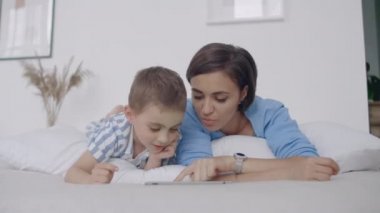 Beyaz yatak odasında, anne ve oğlu tablet ekrana bakmak ve gülmek. Sabah yatakta mutlu aile bir kitap okuma ve bilgisayarın Touchpad tıklayarak