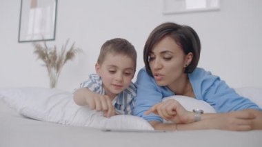 Anne ve oğul beyaz bir yatakta yatan tablet ekrana bakıyor. Tablet bilgisayarınızda oğlunuzla oyun oynayın ve komik videolar izleyin