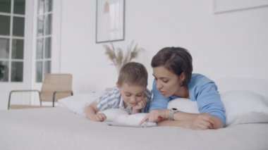 Beyaz yatak odasında, anne ve oğlu tablet ekrana bakmak ve gülmek. Sabah yatakta mutlu aile bir kitap okuma ve bilgisayarın Touchpad tıklayarak