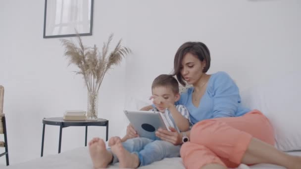 En el dormitorio blanco, mamá e hijo miran la pantalla de la tableta y se ríen. Familia feliz en la cama por la mañana leyendo un libro y haciendo clic en el panel táctil de la computadora — Vídeos de Stock