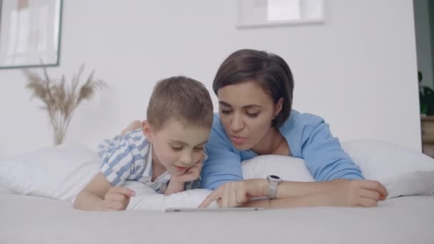 No quarto branco, mãe e filho olham para a tela do tablet e riem. Família feliz na cama pela manhã lendo um livro e clicando no touchpad do computador — Vídeo de Stock