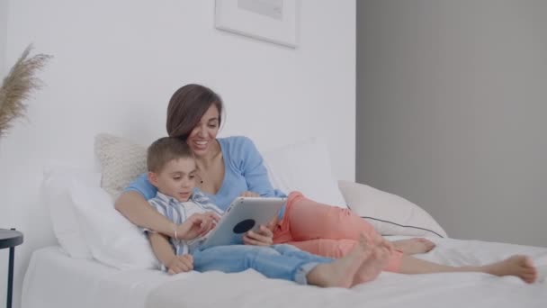 Mamá e hijo mirando la pantalla de la tableta acostados en una cama blanca. Jugar juegos con su hijo en su computadora tableta y ver videos divertidos — Vídeo de stock