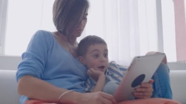 Mutlu bir aile anne ve çocuk oğlu kanepe üzerinde oturan dijital tablet kullanarak, çocuk oğlu ile gülümseyen ebeveyn anne ekran seyir PC bilgisayar online alışveriş yapmak video çağrı izlemek çizgi film yapmak.