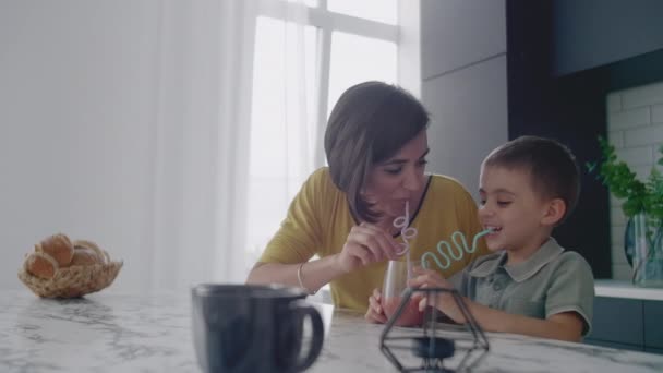 Young mom and son drinking juice with candy at kitchen. Happy mother spending time with son sitting on table in slow motion. — Stock Video