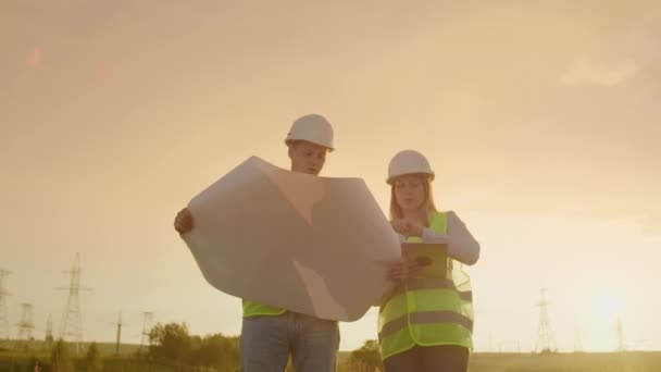 Δύο μηχανικοί, ένας άντρας και μια γυναίκα με κράνη με ένα tablet μηχανικού περπατούν στο χωράφι με πύργους ηλεκτρισμού και συζητούν για την περαιτέρω κατασκευή των Πυργών. — Αρχείο Βίντεο