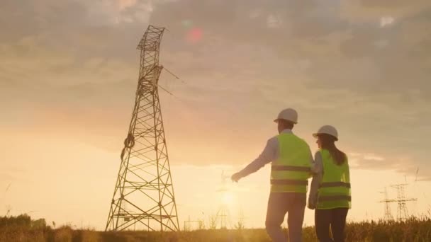 Het uitzicht vanaf de achterkant: twee ingenieurs, een man en een vrouw in helmen met een Tablet van ingenieur lopen op het veld met elektriciteits torens en bespreken de verdere bouw van torens. — Stockvideo