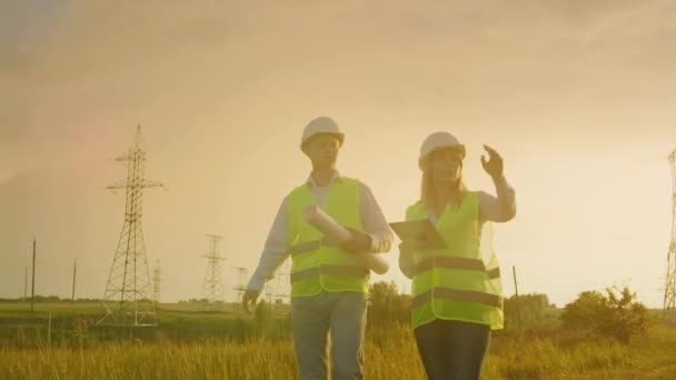 Deux électriciens travaillent ensemble, debout dans le champ près de la ligne de transport d'électricité dans les casques. Debout dans le champ proche avec transmission de puissance — Video