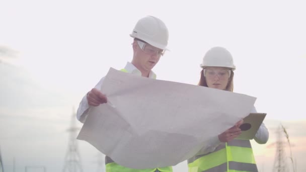 Un groupe d'ingénieurs dans une centrale électrique à haute tension avec une tablette et des dessins marchent et discutent d'un plan pour l'approvisionnement en électricité de la ville. Transport d'énergies propres renouvelables — Video