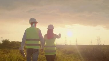 Arkadan görünüm: bir tablet ve çizimler ile yüksek voltajlı bir elektrik santralinde mühendis grubu yürümek ve şehre elektrik arzı için bir plan tartışmak.