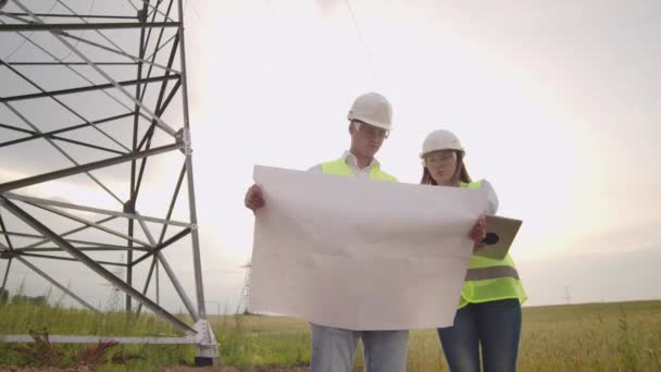 Dos ingenieros, un hombre y una mujer en cascos con una tableta de ingeniero caminan sobre el terreno con torres eléctricas y discuten la construcción de torres . — Vídeo de stock