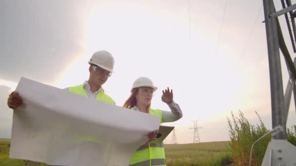 Due elettricisti lavorano insieme, in piedi nel campo vicino alla linea di trasmissione dell'elettricità nei caschi. In piedi nel campo vicino con trasmissione di potenza — Video Stock