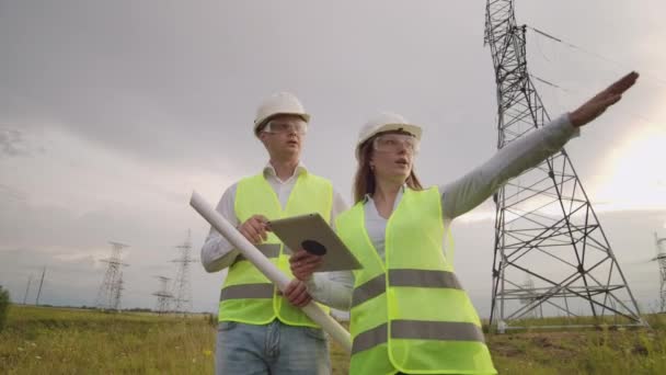 Dwaj Elektrycy współpracują ze sobą, stojąc na polu w pobliżu linii przesyłowych energii elektrycznej w kaskach. Stojąc w polu obok z transmisją mocy — Wideo stockowe