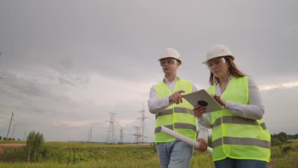 送電線の近くの畑に電気技師の男女。彼は送電線を立てるプロセスを管理する電気技師です. — ストック動画