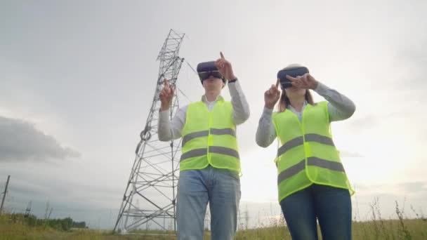 Um homem e uma mulher engenheira em óculos VR controlam a distribuição de energia das redes elétricas e a entrega de eletricidade no fundo de torres elétricas com cabos de alta tensão . — Vídeo de Stock