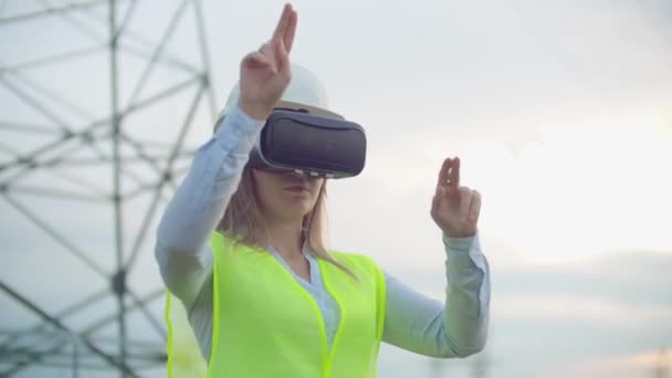 En kvinna elektriker i Virtual Reality glasögon flyttar sin hand simulera arbetet med det grafiska gränssnittet i ett kraftverk mot bakgrund av hög spänning elektriska ledningar — Stockvideo