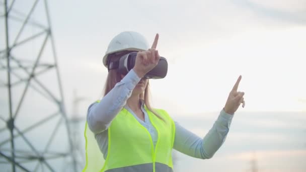 Ingeniera energética femenina de planta media en gafas de realidad virtual y casco blanco en el fondo de torres de línea de alta tensión . — Vídeos de Stock