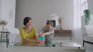 Anne ve oğlu masada oynarken yerde oturan ve elastik bir oyuncak kullanarak gülüyor