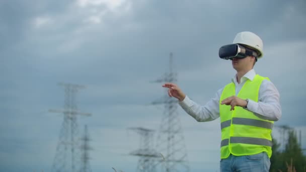 高圧送電線塔を背景にバーチャルリアリティメガネとホワイトヘルメットで中間計画男性エネルギーエンジニア — ストック動画