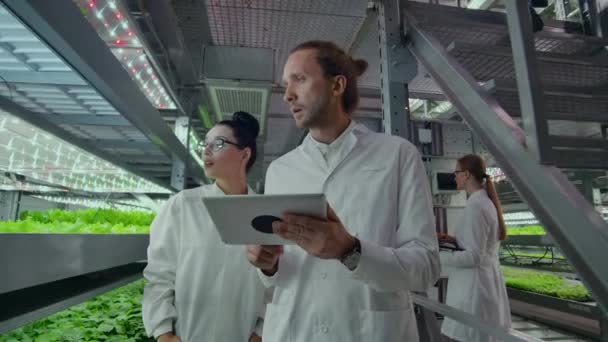 Microbiología en batas blancas ir en una granja moderna con ordenadores portátiles y tabletas estudiando los resultados de las pruebas y la recogida de muestras para el estudio de nuevas especies de plantas . — Vídeo de stock