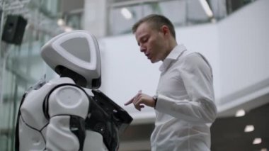 Bir adam bir robot bot ile duruyor ve ona sorular sorar ve robot gövdesi üzerinde ekrana tıklayarak yardım ister.