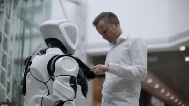 Un hombre con camisa se comunica con un robot blanco haciendo preguntas y presionando la pantalla con los dedos . — Vídeo de stock