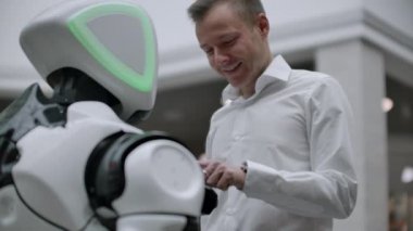 Gömlekli bir adam beyaz bir robotla iletişim kurar ve parmaklarıyla ekrana basarak sorular sorar..