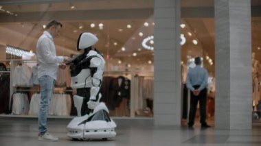 Alışveriş merkezindeki adam bir robot danışmanla iletişim kuruyor. Modern dükkan ve robot satıcısı. Robot Alışveriş Merkezi'nde bir adam yardımcı olur