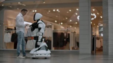 Gömlekli bir adam beyaz bir robotla iletişim kurar ve parmaklarıyla ekrana basarak sorular sorar..