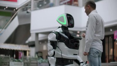 Bir adam bir robot bot ile duruyor ve ona sorular sorar ve robot gövdesi üzerinde ekrana tıklayarak yardım ister.