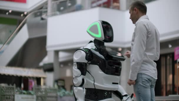 Un homme se tient debout avec un robot robot et lui pose des questions et demande de l'aide en cliquant sur l'écran sur le corps du robot . — Video