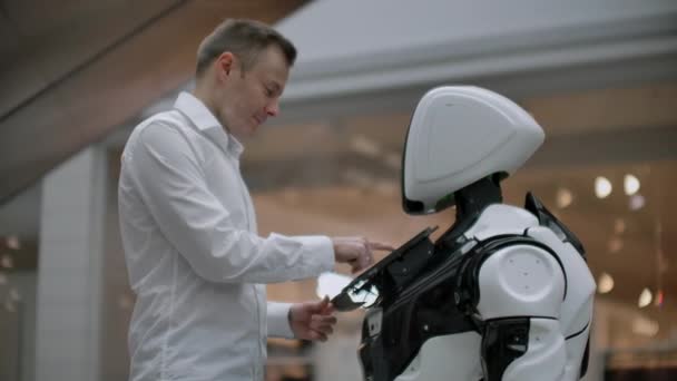 Un homme se tient debout avec un robot robot et lui pose des questions et demande de l'aide en cliquant sur l'écran sur le corps du robot . — Video